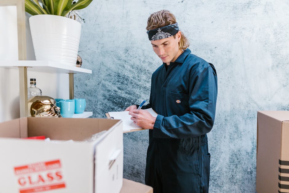 A young male mover wearing a navy-blue uniform and a black bandana with white patterns on his forehead is inside a room, writing on a clipboard. The background features a textured light blue wall and a white shelf holding a large white potted plant, a glass bottle, and blue ceramic cups. In front of him are cardboard moving boxes, one open and partially visible, with packing materials inside. The room appears to be part of a home or storage area, with natural light illuminating the scene. The mover is engaged in organizing or recording details related to a home relocation process, which involves packing and preparing furniture and household items for transport, supported by the presence of packed boxes and the indoor setting.