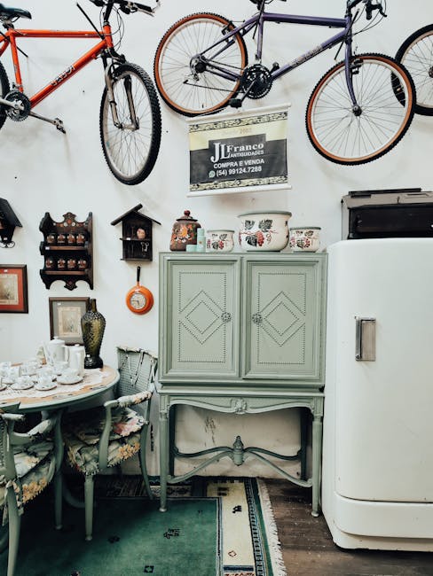 The image depicts a corner of a room with a mix of vintage and modern furnishings. Mounted on the white wall are several bicycles, including a purple bicycle with orange details, a red bicycle, and another with brown tires, all hung by their frames. Below the bikes, a sign with contact information for Franco Autoauffundos is displayed. In the foreground, there is a dining table set with various dishes, cups, and decorative items, accompanied by four upholstered chairs with floral fabric seats. To the right of the table, a light green vintage cupboard with decorative patterns on its doors is situated, with three ceramic jars and a small decorative vase placed on top. Adjacent to the cupboard, a white modern refrigerator is visible. The room features a wooden floor with a small patterned rug partially underneath the table and chairs. Overall, the scene captures a well-organized interior that combines antique and contemporary elements, with an emphasis on home interiors and furniture arrangements that could be related to a house moving or packing process.