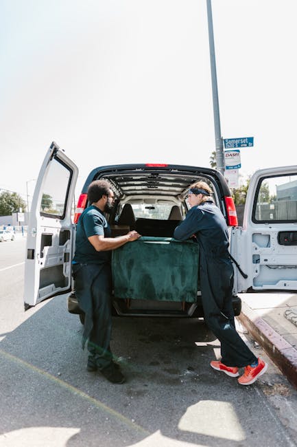A man wearing a blue jacket and dark trousers is seen from behind as he loads a large, white cardboard box into the back of a commercial van parked on a city street. The van's rear doors are open, rev