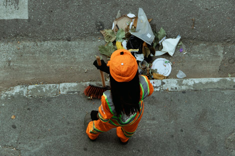 A woman wearing an orange high-visibility jacket with reflective stripes, matching trousers, and an orange cap, is seen from above standing on a paved sidewalk beside a curb. She is holding a broom in