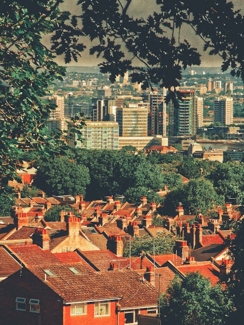 A panoramic view of a suburban area with numerous red-brick houses topped with chimney stacks, surrounded by green trees. The scene is framed by leafy branches in the foreground, and beyond the residential area, a dense cluster of high-rise office buildings and modern skyscrapers extends into the distance under a cloudy sky. The lighting suggests daytime, with natural sunlight illuminating the rooftops and buildings. This outdoor urban landscape captures the contrast between the immediate residential environment and the distant city skyline, reflecting a typical scene in a London suburb. The image emphasizes the closeness of residential homes to central city developments, suitable for illustrating house moves, property relocations, or urban expansion related to [DOMAIN].