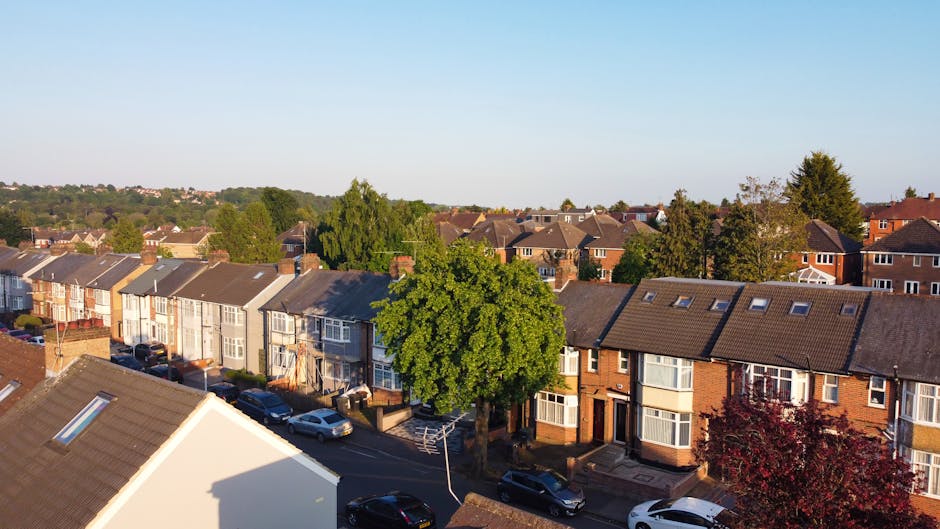 A black and white aerial photograph showing a residential neighbourhood with rows of terraced houses, detached and semi-detached homes, and tree-lined streets. The scene includes a perspective view of rooftops, front gardens, and parked cars along the road. The image captures a wide expanse of the suburb with a distant city skyline visible on the horizon under partly cloudy skies. In the foreground, several houses display yards with garden furniture, fences, and rectangular driveways. The streets are lined with vehicles, and some homes have visible porches or carports. The overall environment suggests a quiet, established area suitable for home relocation and furniture transport services. This context aligns with the services offered by Man with Van Winchmore Hill for house removals and packing and moving logistics within the N21 postcode area.