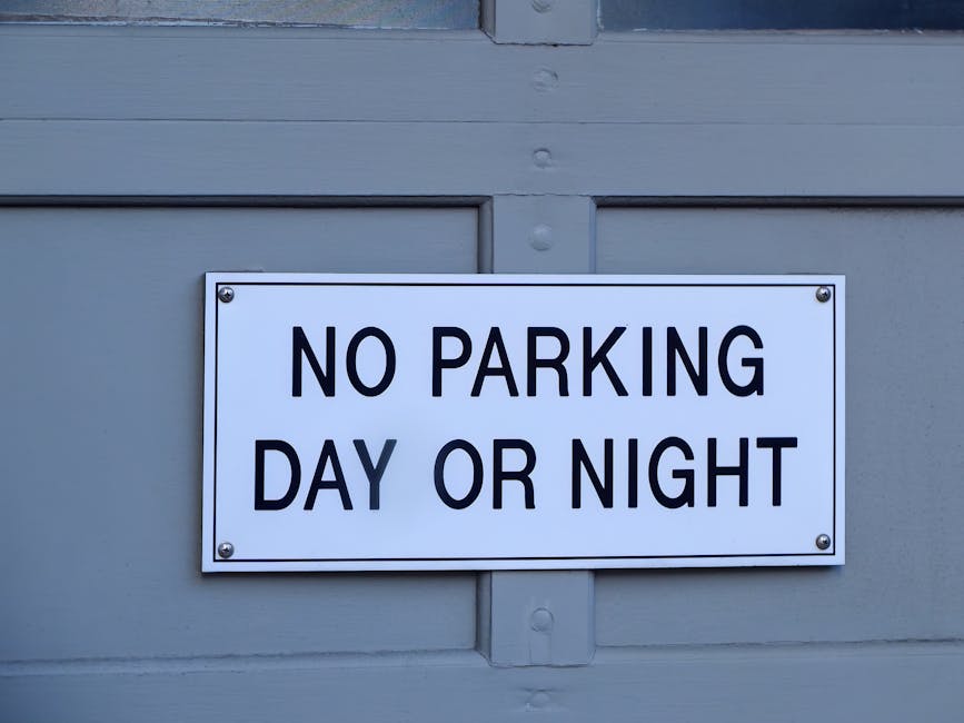 A rectangular white sign with bold black capital letters reading 'NO PARKING DAY OR NIGHT' mounted on a light grey wooden garage door. The sign is attached with four screws, one in each corner, and positioned centrally on the panel. The garage door consists of horizontal wooden panels, with visible joints and panels above the sign. The scene is well-lit with natural daylight, and the overall setting relates to parking restrictions that may impact house removals or furniture transport in Winchmore Hill, as referenced in the page about permits and parking rules for home relocations. The sign's clear message highlights parking limitations relevant to moving logistics managed by Man with Van Winchmore Hill.