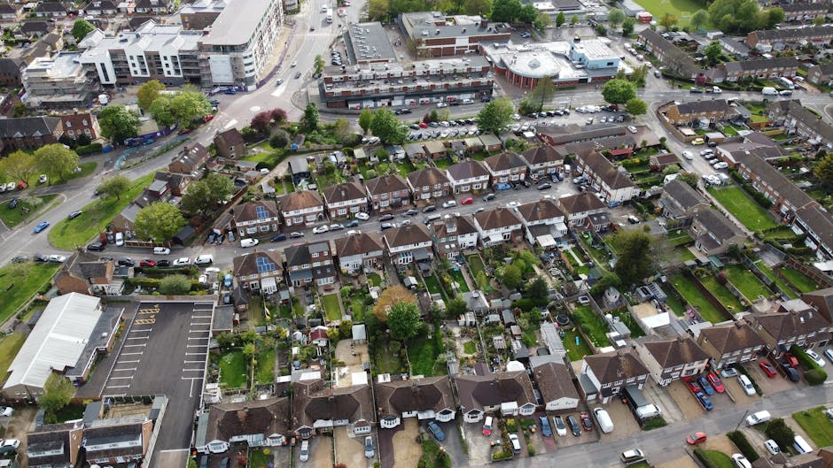 An aerial view of a residential neighbourhood in Winchmore Hill, showing a row of terraced houses with small gardens, some with patio furniture and greenery, and parking spaces with cars along the street. In the background, a commercial area with larger buildings, parking lots, and a busy road is visible. The image captures the typical layout of suburban housing and nearby infrastructure, illustrating the environment where home relocation and furniture transport services by Man with Van Winchmore Hill can be conducted during the loading process for removals and moving logistics.