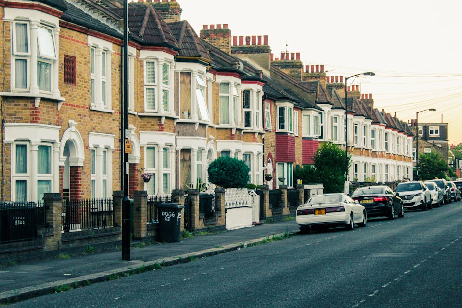 A row of traditional British terraced houses on a quiet residential street in Winchmore Hill, with brick facades, bay windows, and small front gardens enclosed by low brick walls and wrought iron fences. Several parked cars, including sedans and hatchbacks, line the street directly in front of the homes. The pavement features a black lamppost and a black wheelie bin near one of the houses. Street lamps extend along the road, which is shaded with soft evening light, and overhead power lines run parallel to the street. This scene illustrates a typical suburban area often serviced by home relocation and furniture transport companies such as Man with Van Winchmore Hill, highlighting street access that may require permits for moving large items or household furniture during a home removal or packing and moving process.
