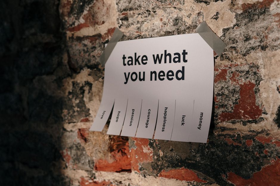 A white sheet of paper taped to a rough brick wall with brown packing tape, displaying the heading 'take what you need' in bold black letters. The paper features several vertical columns with individual words such as 'hope,' 'passion,' 'courage,' 'happiness,' 'luck,' and 'money,' each possibly torn or pulled at the bottom edges, suggesting a shared or communal message. This signage is outside a property, indicating an informal or community-driven initiative related to moving or relocation, possibly encouraging support or sharing resources among people planning home moves. The environment appears outdoors, with natural lighting illuminating the wall and signage, which might be used as a notice or encouragement during the home relocation process facilitated by [COMPANY_NAME], or as part of a broader moving or packing activity near the property.