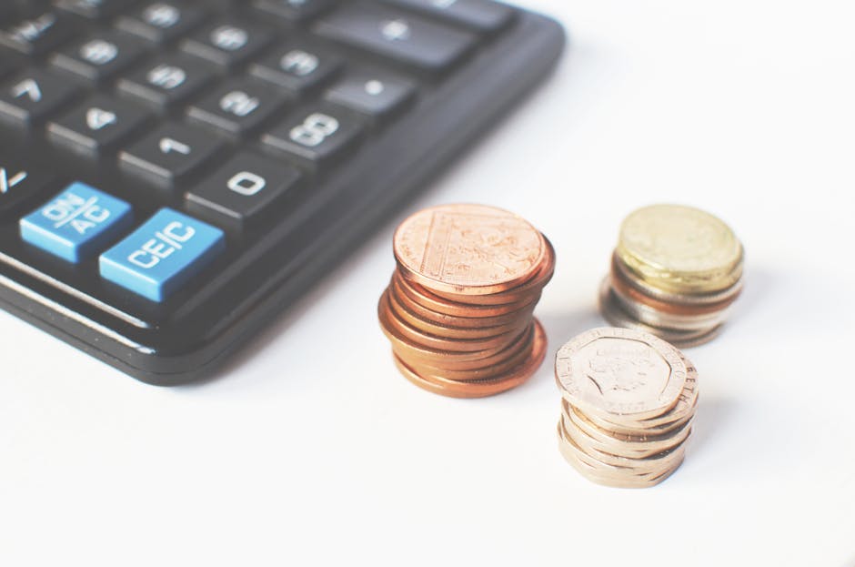 Close-up of a black calculator with blue and white buttons, positioned on a white surface next to several stacks of coins, including copper, nickel, and gold-coloured ones, which are arranged in small piles. The scene suggests a focus on cost or budgeting, relevant to home relocation and moving services offered by Man with Van Winchmore Hill. The coins are placed near the edge of the calculator, indicating financial calculations related to removal estimates, pricing, and moving logistics. The lighting is bright, emphasizing the details of the calculator and coins, while the plain background keeps the focus on the objects. This visual context supports understanding of the costs associated with furniture transport and packing and moving processes, as outlined in the page about understanding removal estimates in Winchmore Hill.
