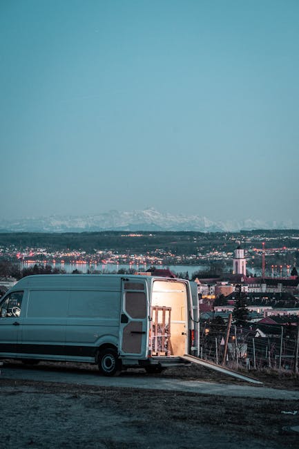 A white moving van parked outdoors on a sloped surface during dusk or early evening, with its rear doors open revealing a wooden ladder and packing supplies inside. The van is positioned in front of a residential area with houses and industrial buildings, and a city skyline can be seen in the distance under a fading blue sky. The scene suggests a home relocation or furniture transport process as part of a house removals service. The area is illuminated by natural light, and the open doors indicate active loading or unloading of moving materials, consistent with professional removals activities. This setting aligns with the services offered by Man with Van Winchmore Hill, specializing in urgent or last-minute moves, with a focus on efficient loading and transport logistics during evening hours.