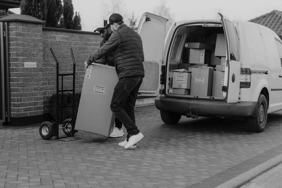 A white moving van parked outdoors on a sloped surface during dusk or early evening, with its rear doors open revealing a wooden ladder and packing supplies inside. The van is positioned in front of a residential area with houses and industrial buildings, and a city skyline can be seen in the distance under a fading blue sky. The scene suggests a home relocation or furniture transport process as part of a house removals service. The area is illuminated by natural light, and the open doors indicate active loading or unloading of moving materials, consistent with professional removals activities. This setting aligns with the services offered by Man with Van Winchmore Hill, specializing in urgent or last-minute moves, with a focus on efficient loading and transport logistics during evening hours.