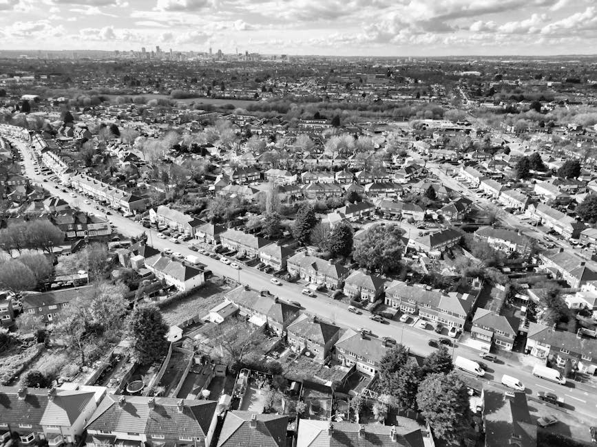 An aerial black-and-white photograph of a residential neighbourhood showing rows of terraced houses with pitched roofs, some with small gardens or backyards, and a main street with parked cars along both sides. The street runs through the middle of the image, with additional houses extending into the distance. Trees and greenery are visible throughout the area, with a broader suburban landscape and a distant city skyline featuring high-rise buildings on the horizon. This view captures the layout of the houses, streets, and surrounding environment typical for a home relocation or moving logistics context, reflecting the urban spread and community setting for furniture transport or packing and moving services by Man with Van Winchmore Hill.